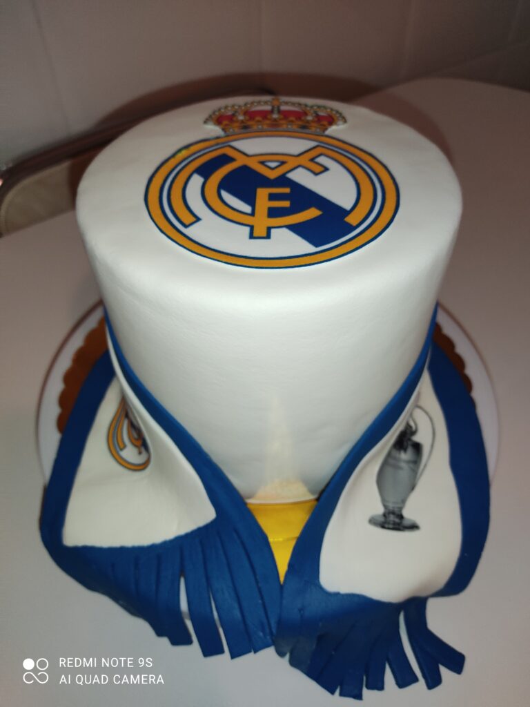 Tarta personalizada de fondant del Real Madrid con escudo, bufandas y temática de fútbol, ideal para cumpleaños de fans merengues.